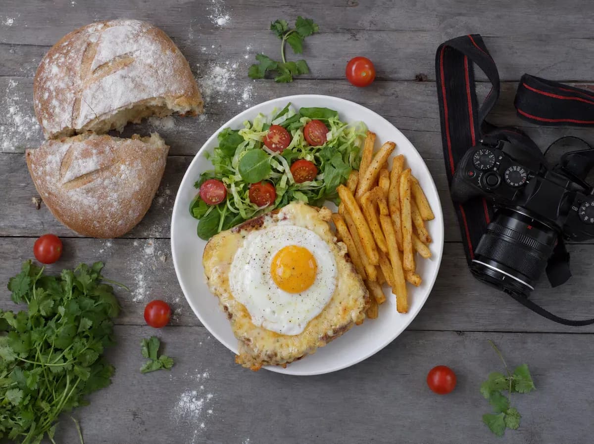 Food and beverage: A top-down food scene on weathered grey planks. A white plate holds a croque-mad