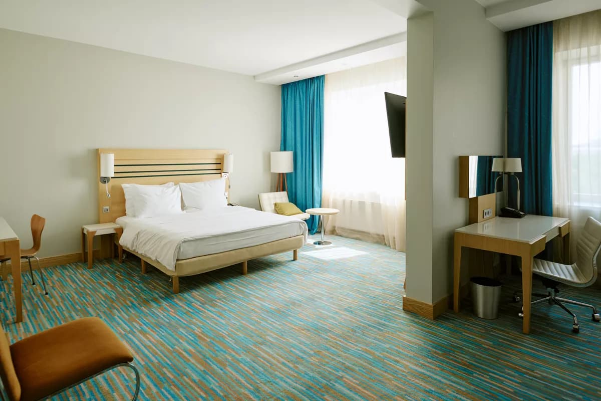 Real photograph — Interior: A spacious modern hotel room. A double bed with a pale wood slatted headboard, c