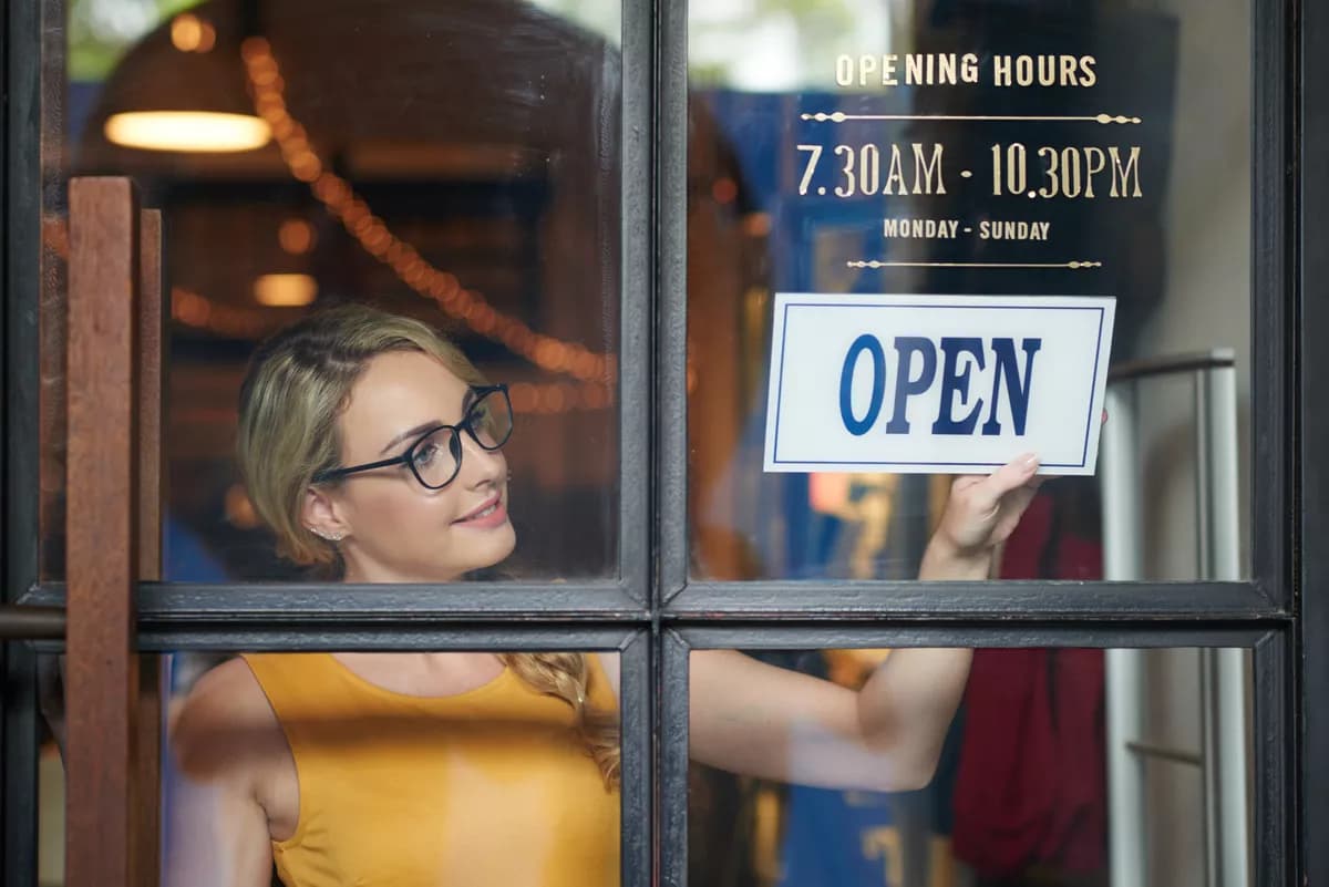 Real photograph — Layout and typography: A woman seen through a shop window as she hangs a sign. She stands inside a diml