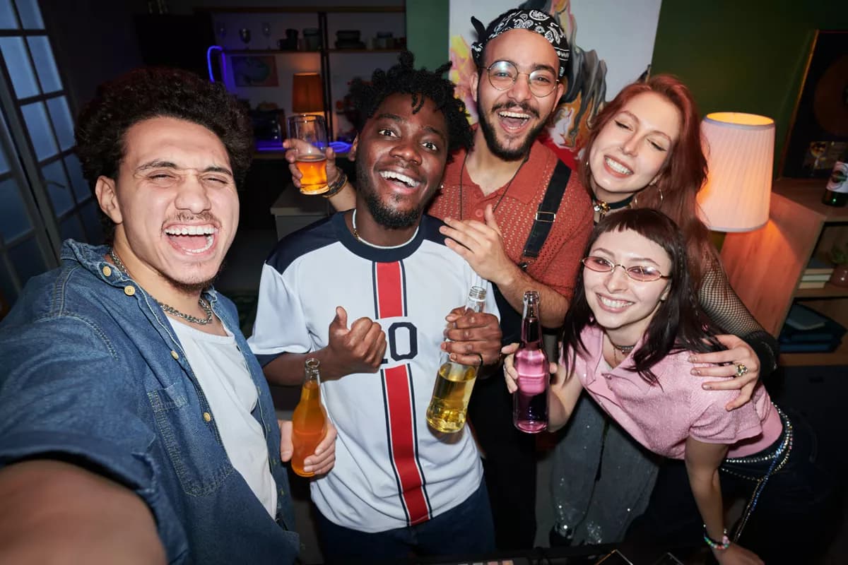 Real photograph — People and lifestyle: A wide-angle group selfie of five friends at a house party, all laughing and cro